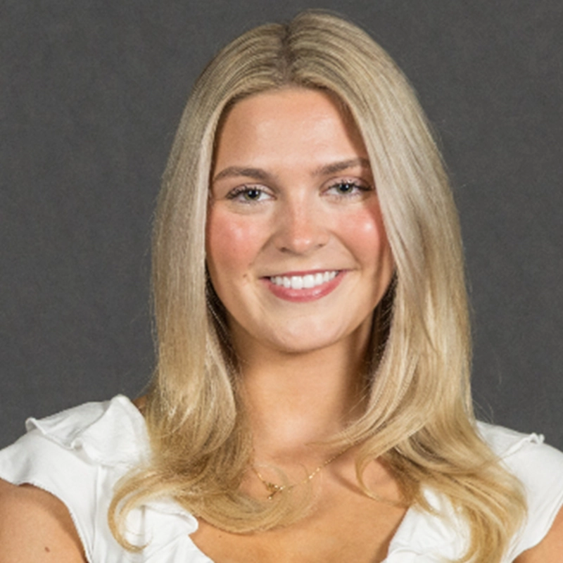 Addison Wivagg, a young woman with long, straight blonde hair, smiles at the camera. She is wearing a white blouse with ruffled sleeves and a delicate gold necklace. The plain, dark gray background highlights her friendly expression.