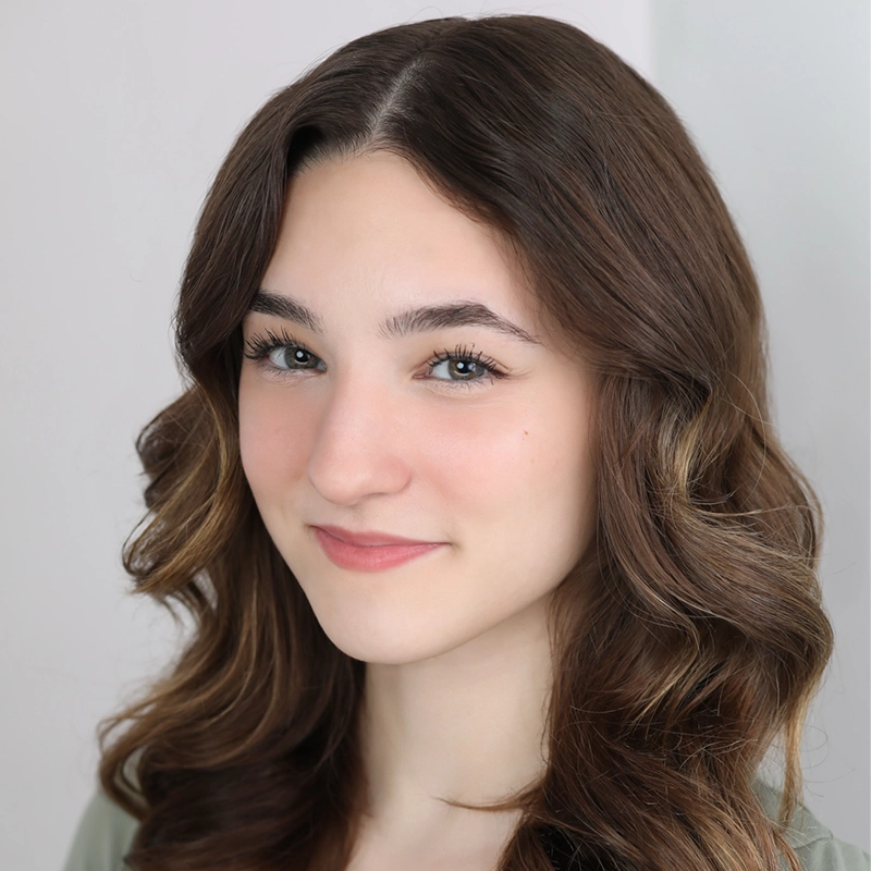 A young woman, Shayla Lopez, with long, wavy brown hair and fair skin smiles softly at the camera. She has light blue eyes, subtle makeup, and a pale green top against a plain white background that keeps the focus on her face.