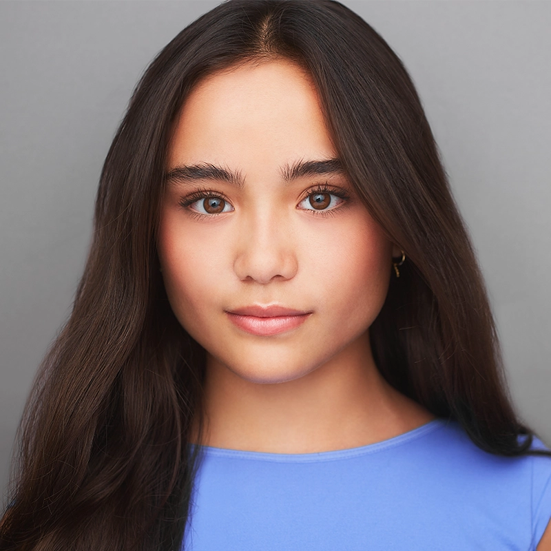 A young woman with long, straight dark brown hair and clear skin looks directly at the camera. She wears a light blue top and small gold hoop earrings. The background is plain gray, and she has a neutral, calm expression.