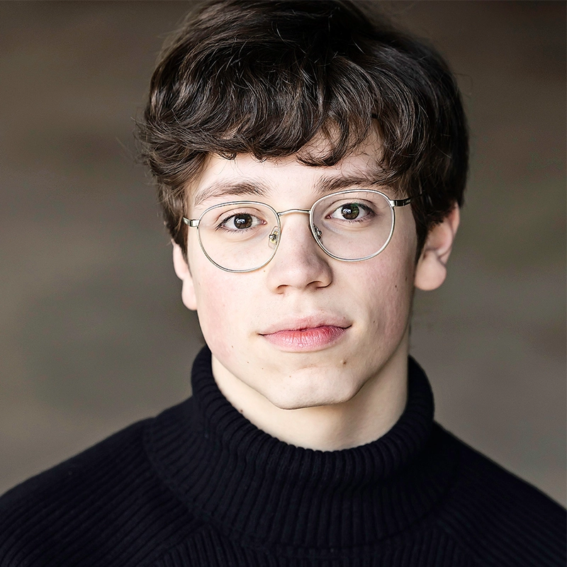Shayla Lopez, a young person with short, wavy brown hair and round eyeglasses, looks directly at the camera. They wear a black, ribbed turtleneck sweater against a blurred, neutral background that draws focus to their face.