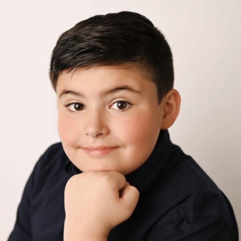 A young boy with short dark hair and fair skin smiles softly at the camera. He is wearing a dark shirt and rests his chin on his hand in a relaxed pose against a plain, light-colored background.