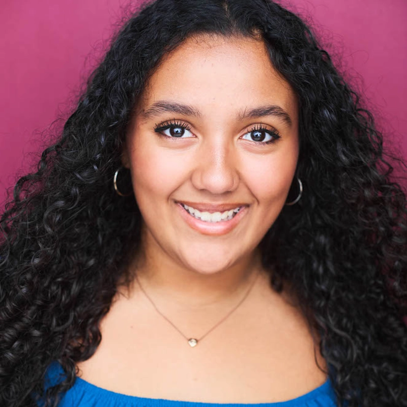 Sophia Leverett, a young woman with long curly dark hair, smiles at the camera. She wears gold hoop earrings, a delicate necklace, and an off-the-shoulder blue top. The solid magenta background highlights her warm skin and bright eyes.