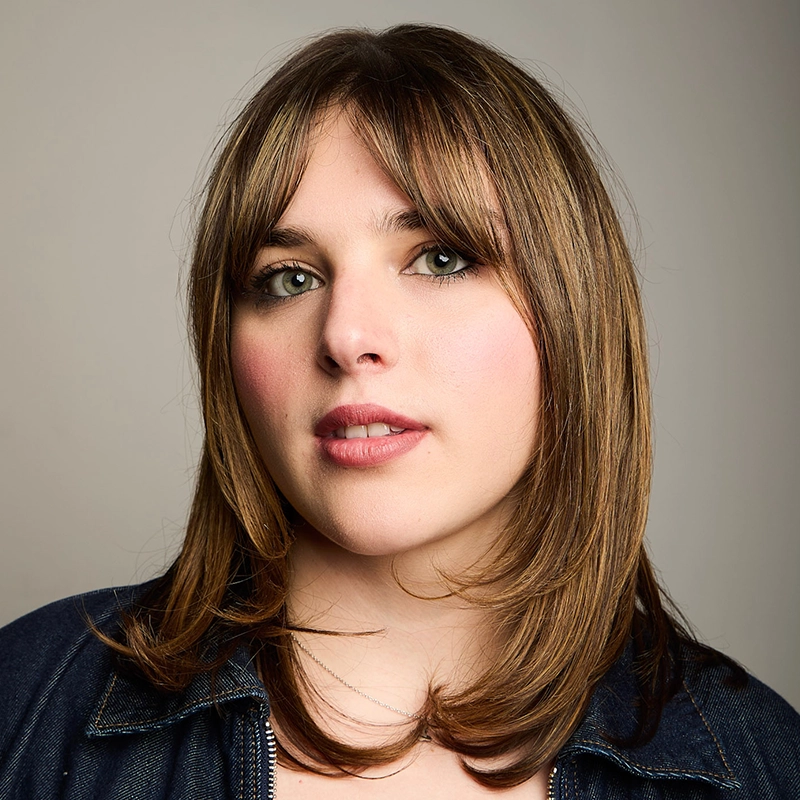 A young woman with straight, light brown hair and bangs looks at the camera. She has light skin, green eyes, natural makeup, and wears a denim jacket. The softly lit background completes this portrait of Alice K. Tinari.