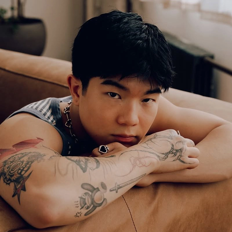 Aedan Alvarez, a young person with short black hair, leans on a tan couch gazing at the camera. Tattoos of a cartoonish bear and dragon decorate their arms. They wear a sleeveless top, chain necklace, and ring as soft indoor light streams in behind them.