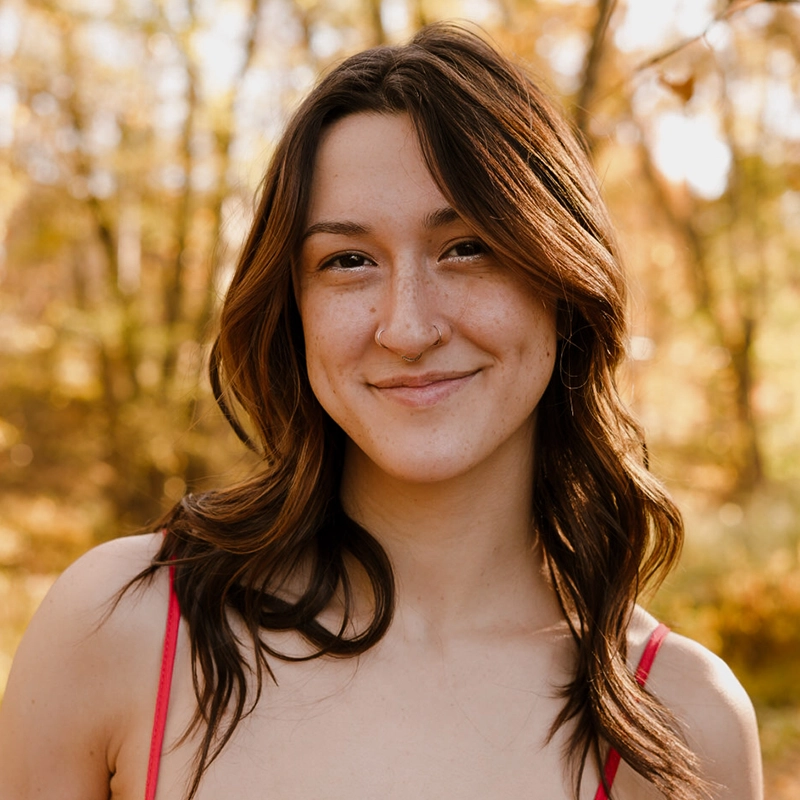 A young woman with wavy brown hair, light skin, and a nose ring smiles gently outdoors. She wears a pink top and stands in front of blurred autumn trees with golden leaves in soft sunlight.