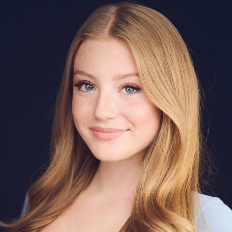 Shayla Lopez, a young woman with long, wavy strawberry-blonde hair and fair skin, smiles softly. Her blue eyes and subtle makeup complement her light blue top, while the dark background highlights her features.