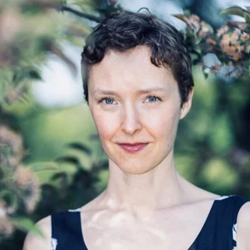 Amanda Quaid, with short curly brown hair and blue eyes, stands outdoors among blurred green foliage, wearing a sleeveless dark top and looking directly at the camera with a slight smile. The lighting is natural and soft.