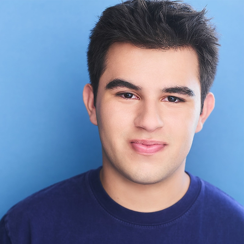 Sophia Leverett, a young person with short dark hair and thick eyebrows, smiles gently at the camera. They are wearing a dark blue crewneck shirt and standing in front of a smooth blue background with bright, even lighting.