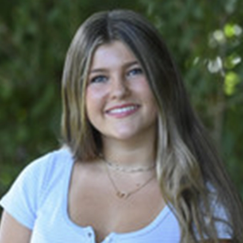 A young woman with long, straight, light brown hair and blue eyes smiles at the camera. Phebe Barnett is wearing a white short-sleeved shirt and two layered necklaces, set against a backdrop of blurred greenery in an outdoor setting.