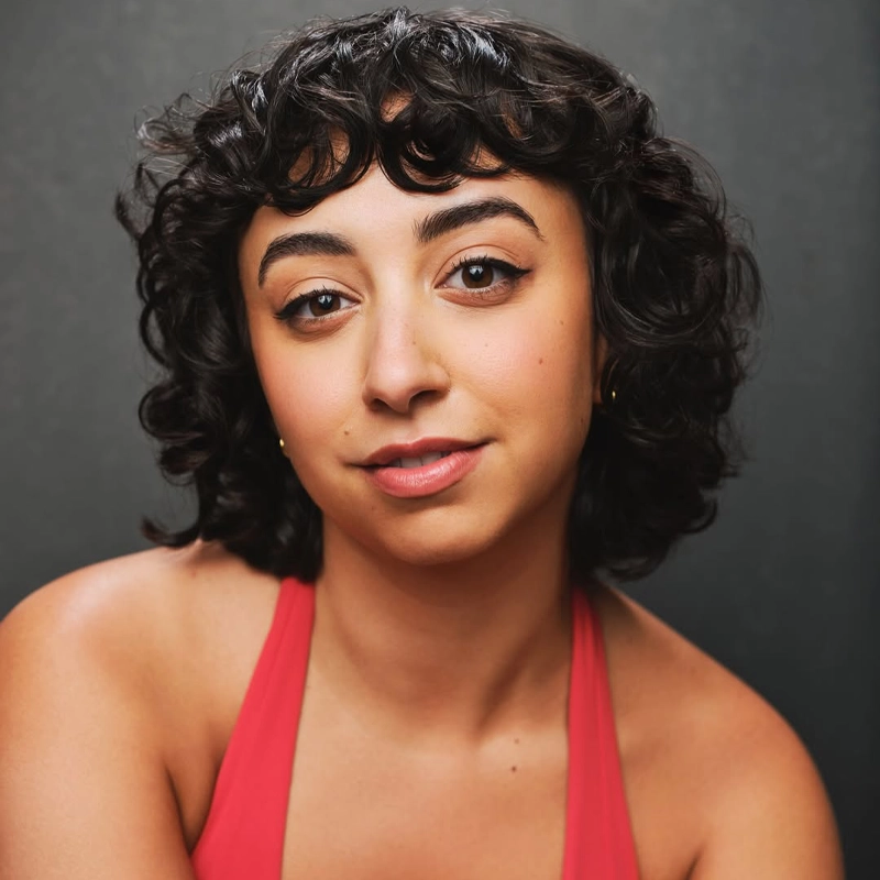A young woman with short, curly dark hair and winged eyeliner gazes softly at the camera. She wears a sleeveless red top and gold hoop earrings. The simple, dark gray background creates an intimate portrait of Caro Daye Attayek.