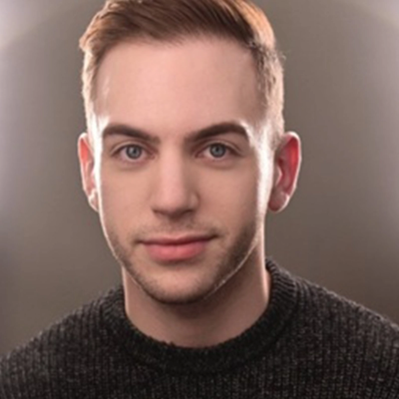 A young man with fair skin, short light brown hair, and blue eyes looks at the camera. He is wearing a dark, textured sweater. The softly blurred background with smooth lighting highlights his face, reminiscent of an Ellie Fishman portrait style.