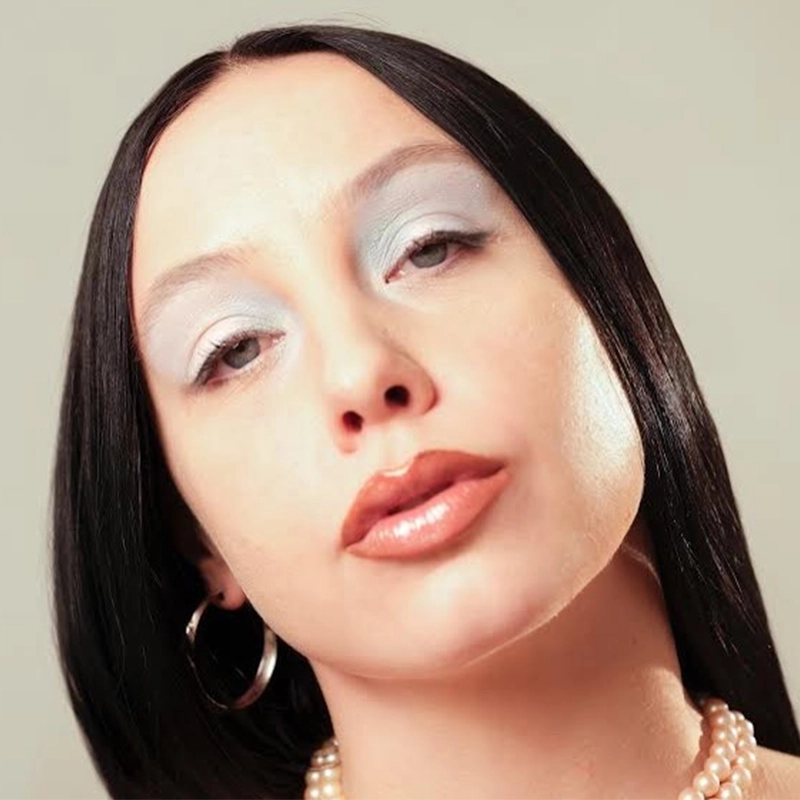 A person with straight, dark hair parted in the middle gazes at the camera. They wear pearl earrings, a pearl necklace, glossy nude lipstick, and shimmery pale blue eye shadow. The background is plain and softly lit.