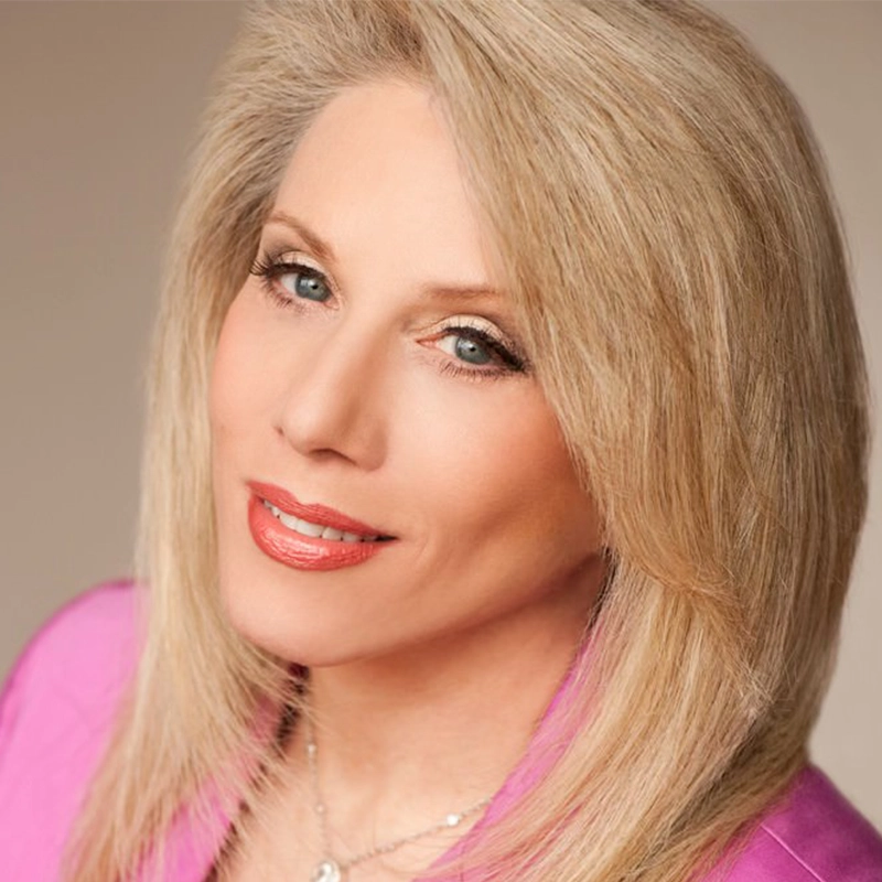 A woman with straight, shoulder-length blonde hair and light blue eyes smiles softly. She is wearing a bright pink blazer and subtle makeup with glossy lipstick. The background is softly blurred in warm tones, keeping the focus on her face.