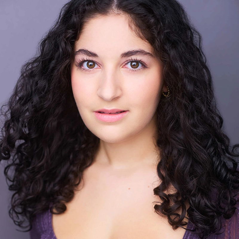 A young woman, Addison Wivagg, with long, dark, curly hair and fair skin looks at the camera. She has hazel eyes, light makeup, and wears a purple top. The background is a plain, soft gray, creating a simple, professional headshot.