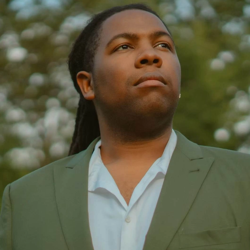 Brandon Bell, with long dark hair and medium-dark skin, wears a light green blazer over an open-collared white shirt, gazing confidently into the distance. The background is blurry, filled with green foliage and bokeh light.