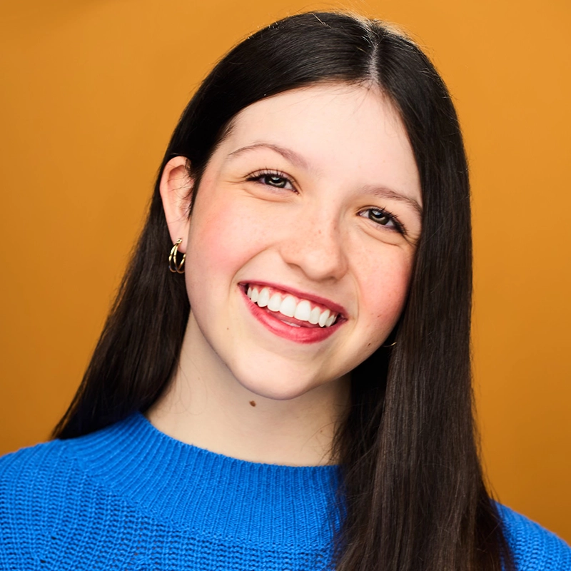 A young woman with straight, long dark hair smiles warmly at the camera. She wears a bright blue sweater and small gold hoop earrings. Her background is a solid, warm orange color, creating a cheerful and inviting portrait.