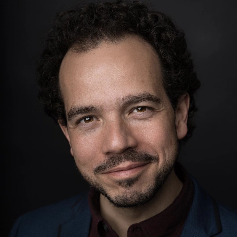 Felipe Bombonato, with short curly dark hair and a trimmed beard, smiles softly. He wears a dark jacket over a burgundy shirt. The black background and warm lighting highlight his approachable expression.