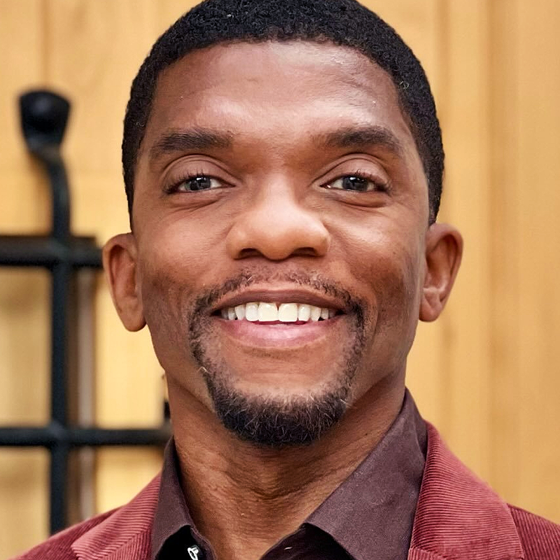A smiling man with short black hair and a goatee, identified as Kevin Boseman, wears a burgundy jacket over a brown shirt. He stands indoors in front of a blurred wooden wall with black metal details. The photo is bright and clear.