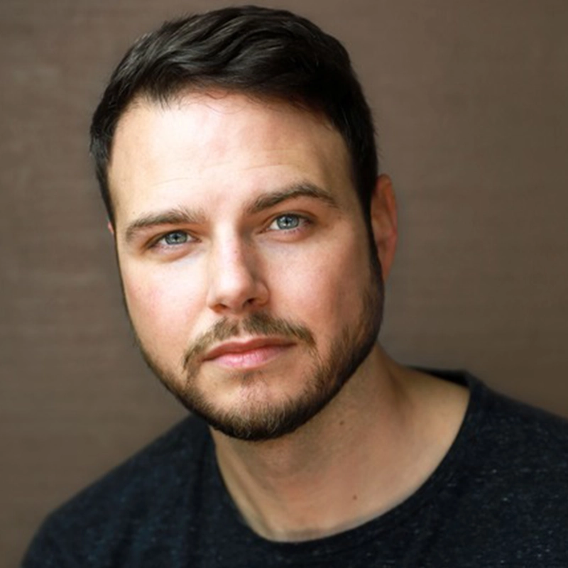 A man with short dark hair, blue eyes, and a trimmed beard gazes softly at the camera. He wears a dark shirt, set against a plain brown background that echoes Ellie Fishman's signature warmth and natural tones.