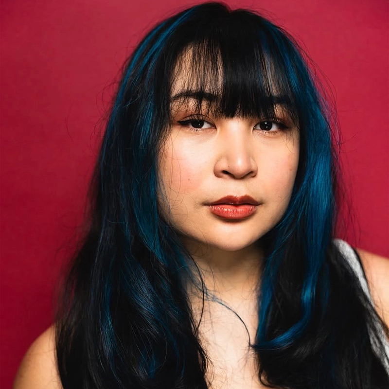 Amaya Braganza, with long black hair featuring blue highlights and straight-cut bangs, poses against a solid fuchsia background. She has natural makeup with winged eyeliner and glossy lips, and wears a sleeveless white top.