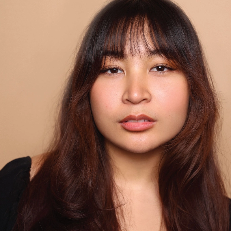 A young woman with long, wavy dark brown hair and bangs poses against a beige background. Amaya Braganza has warm-toned skin, natural makeup, and wears a black top with an off-shoulder sleeve barely visible. She gazes softly at the camera.