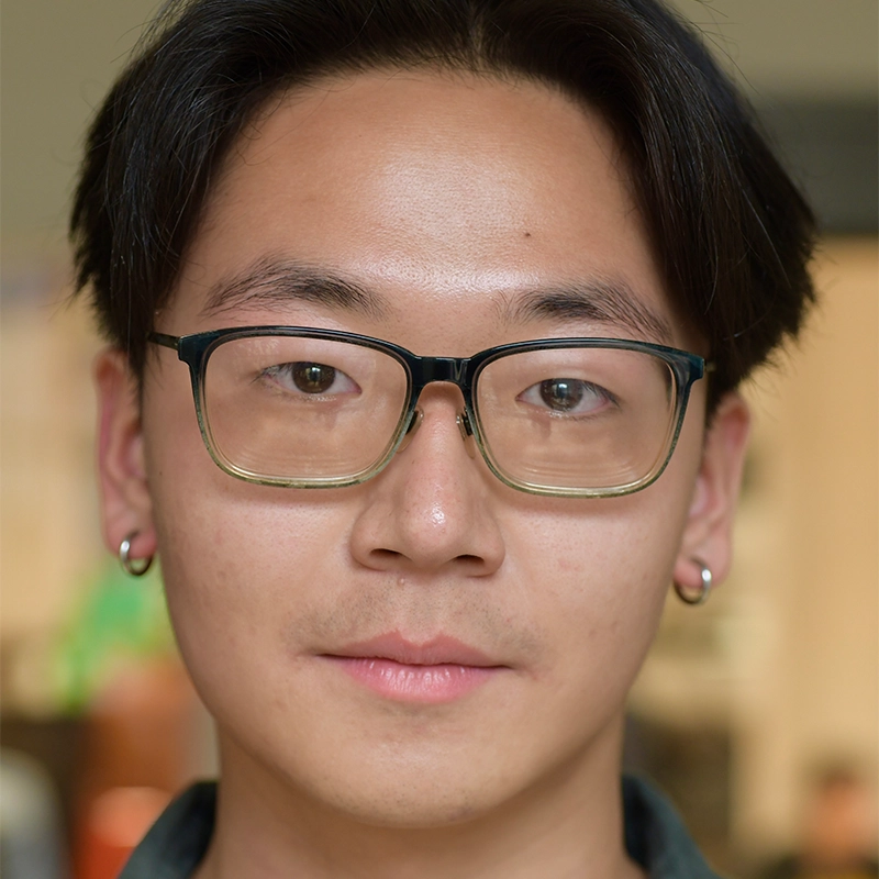 A young person with short, straight dark hair, wearing large, rectangular glasses and small hoop earrings. They have a neutral expression and clear skin. The background is softly blurred, suggesting an indoor setting.