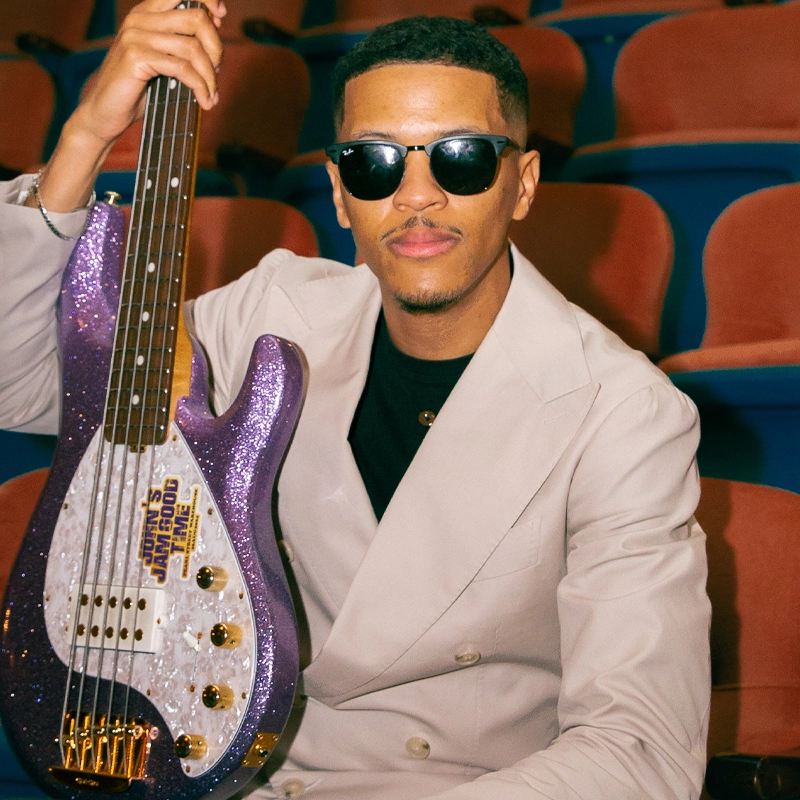 A man wearing dark sunglasses and a light beige suit sits in a theater seat, holding a purple glittery electric bass guitar. The background shows empty, red-upholstered seats. John David Bratton II looks at the camera with a confident expression.