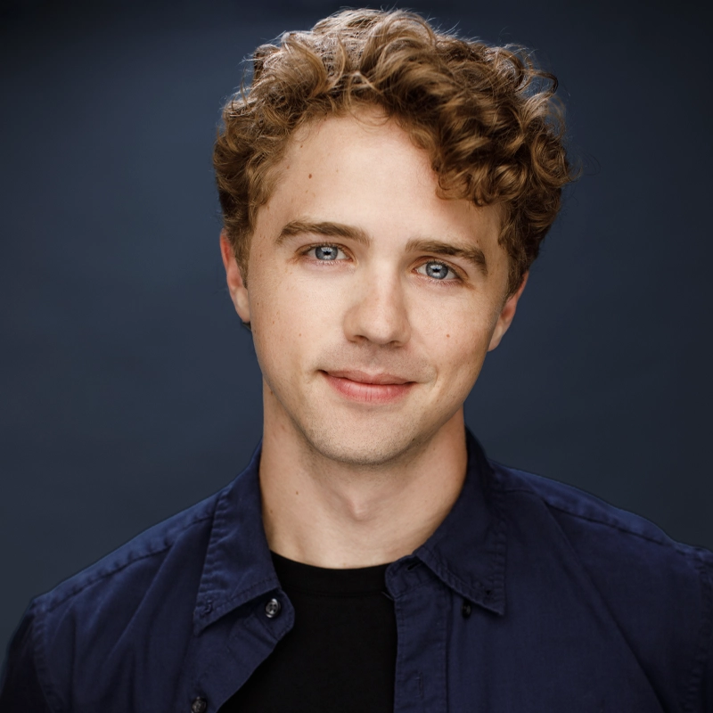 A headshot of Caden Brauch, a young man with wavy, reddish-brown hair and light blue eyes looking directly at the camera. He is wearing a dark blue button-down shirt over a black t-shirt against a simple, dark blue studio background.