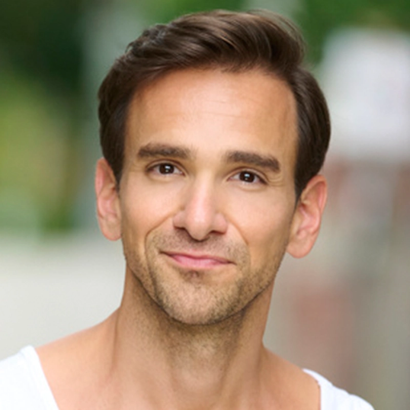 Brent Thiessen, a man with short brown hair and a light beard, smiles softly at the camera. He is wearing a white shirt and standing outdoors, with a blurred green and white background suggesting trees or foliage.