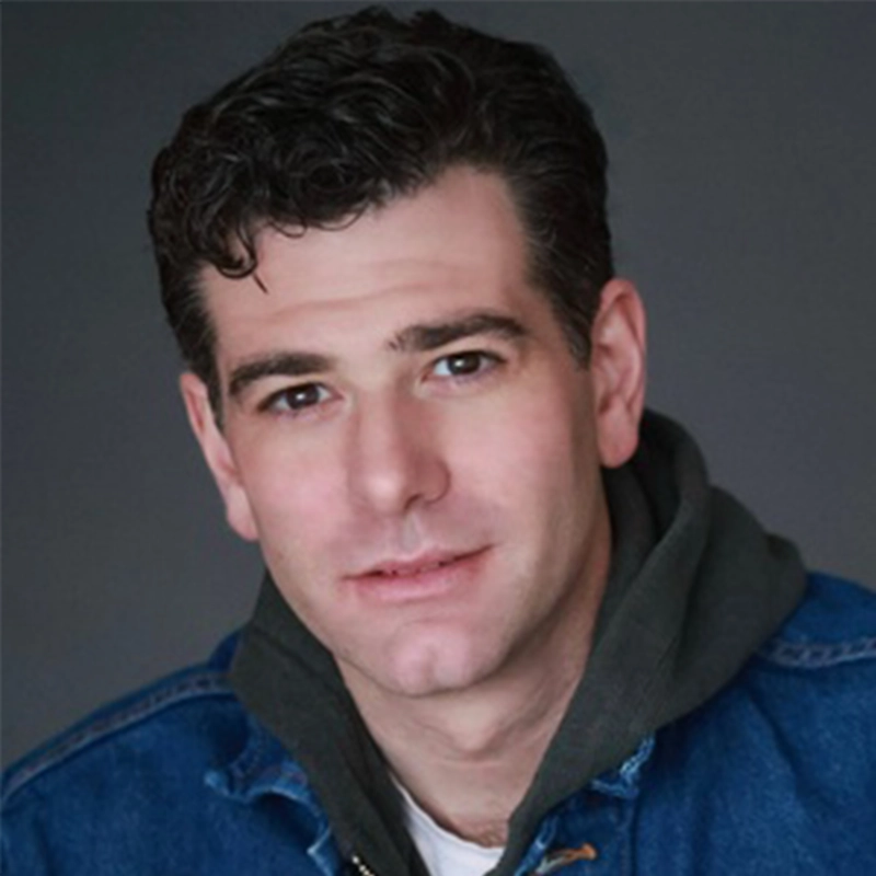A man with short, dark, curly hair and fair skin looks at the camera with a slight smile. He wears a dark hoodie under a blue denim jacket. The plain, dark background creates a professional portrait setting reminiscent of Ellie Fishman's style.