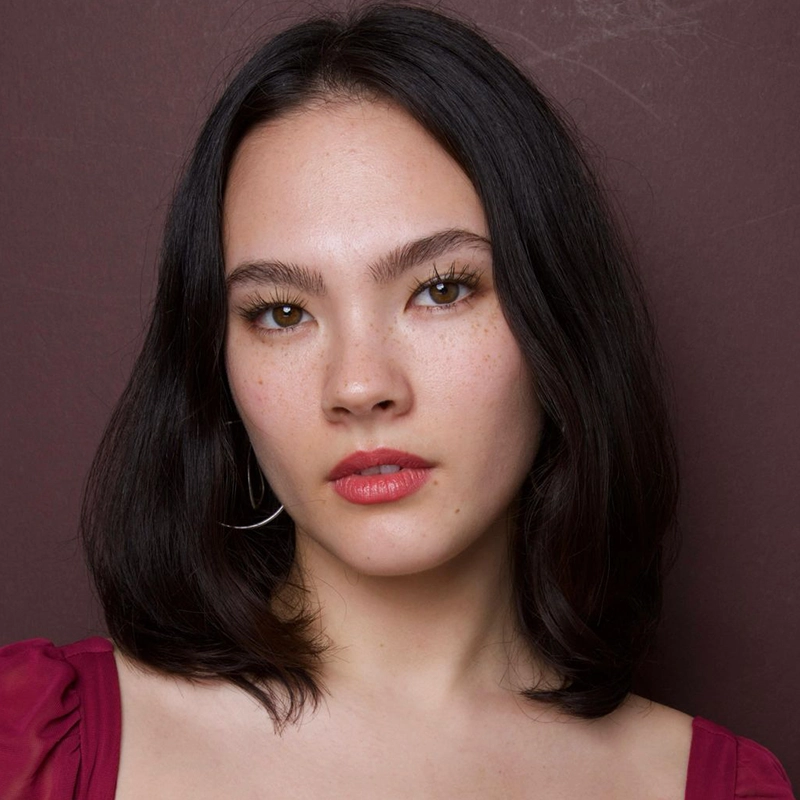 Caitlin Balón, a young woman with shoulder-length dark hair, wears a red top and hoop earrings as she gazes directly at the camera. She has clear, fair skin with freckles, glossy lips, natural makeup, and a neutral expression against a plain burgundy background.