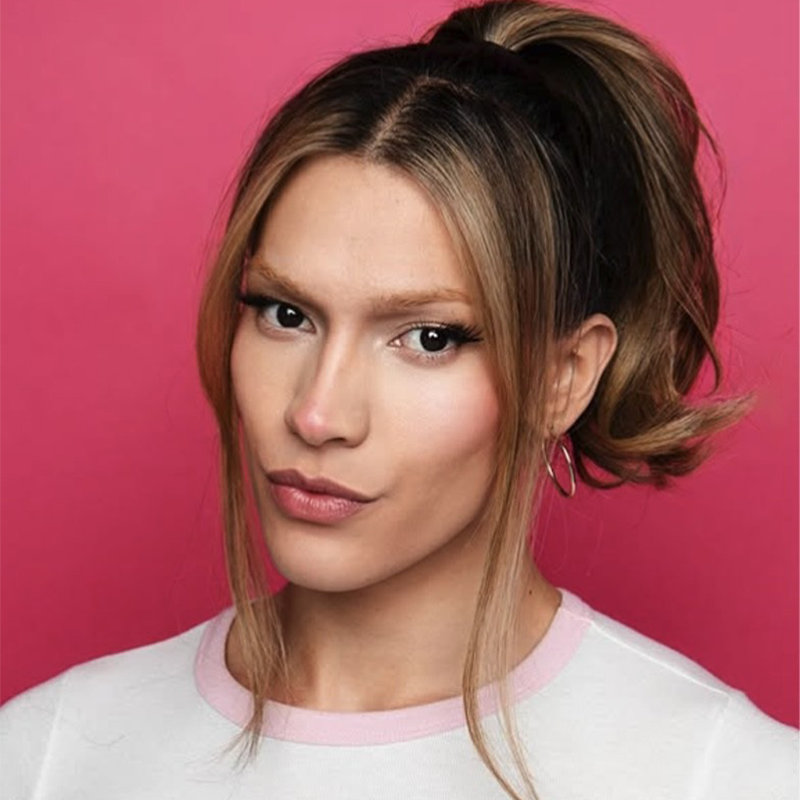 A person with long, highlighted hair styled in a high ponytail with loose strands framing their face poses in front of a solid pink background. Inspired by Caleb Quezon, they wear a white top with pink trim and small hoop earrings.