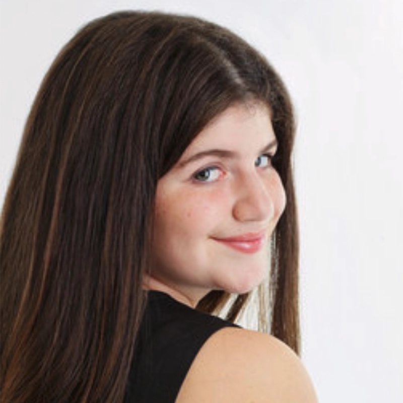 A young person with long, straight brown hair is turned slightly over their shoulder, smiling softly at the camera. They are wearing a sleeveless black top, and the plain, light background draws attention to their gentle expression—perhaps capturing a candid moment reminiscent of Timothy Thomas’s understated style.