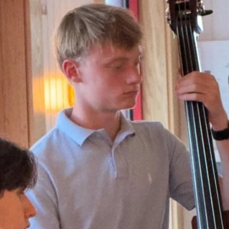 A young man with short blond hair, wearing a light blue polo shirt, plays an upright bass indoors. He is focused on the instrument, hands expertly placed, as warm lighting and wooden walls surround William Calohan in the background.