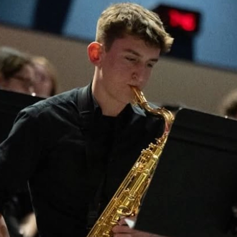 Chase Cassidy, a young person with short brown hair in a black shirt, plays an alto saxophone. The background is out of focus, showing other musicians and blue walls, suggesting a performance or rehearsal setting.