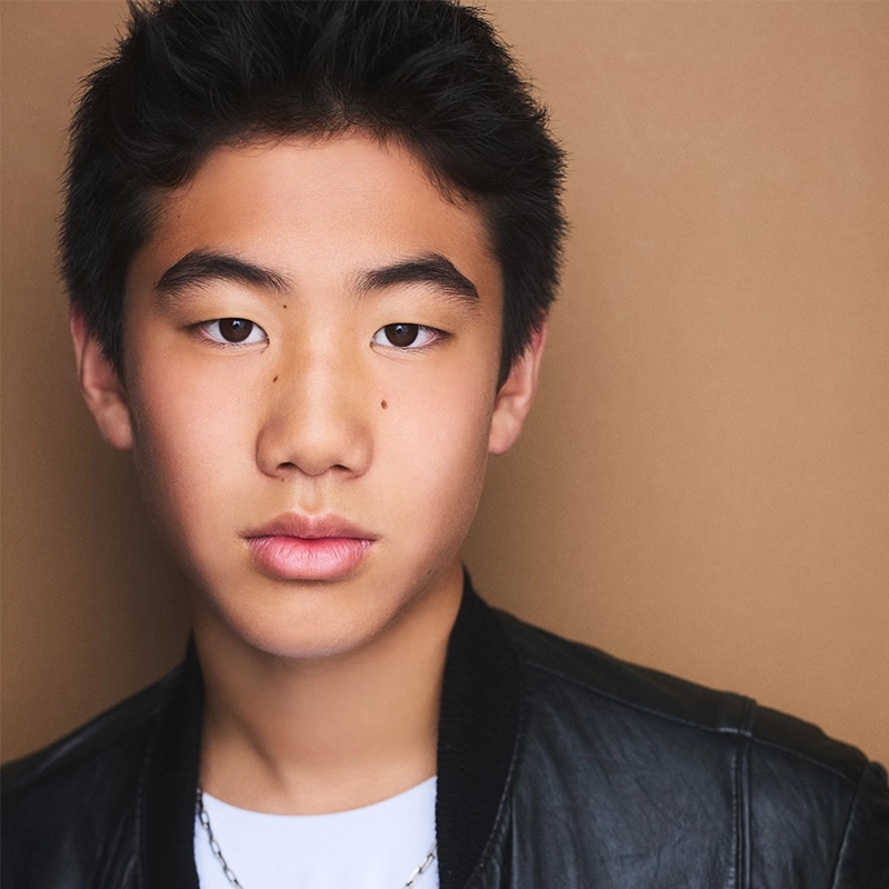 A teenage boy with short black hair and smooth skin stares seriously at the camera. He wears a black leather jacket over a white shirt and necklace. The background is plain and light brown, creating a neutral setting for his portrait.