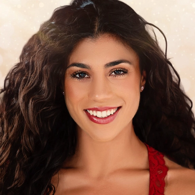 Lauren Celentano, with long, dark curly hair and blue eyes, smiles brightly. She wears a red lace top and small stud earrings. The softly blurred background with light bokeh creates a warm, glowing atmosphere.