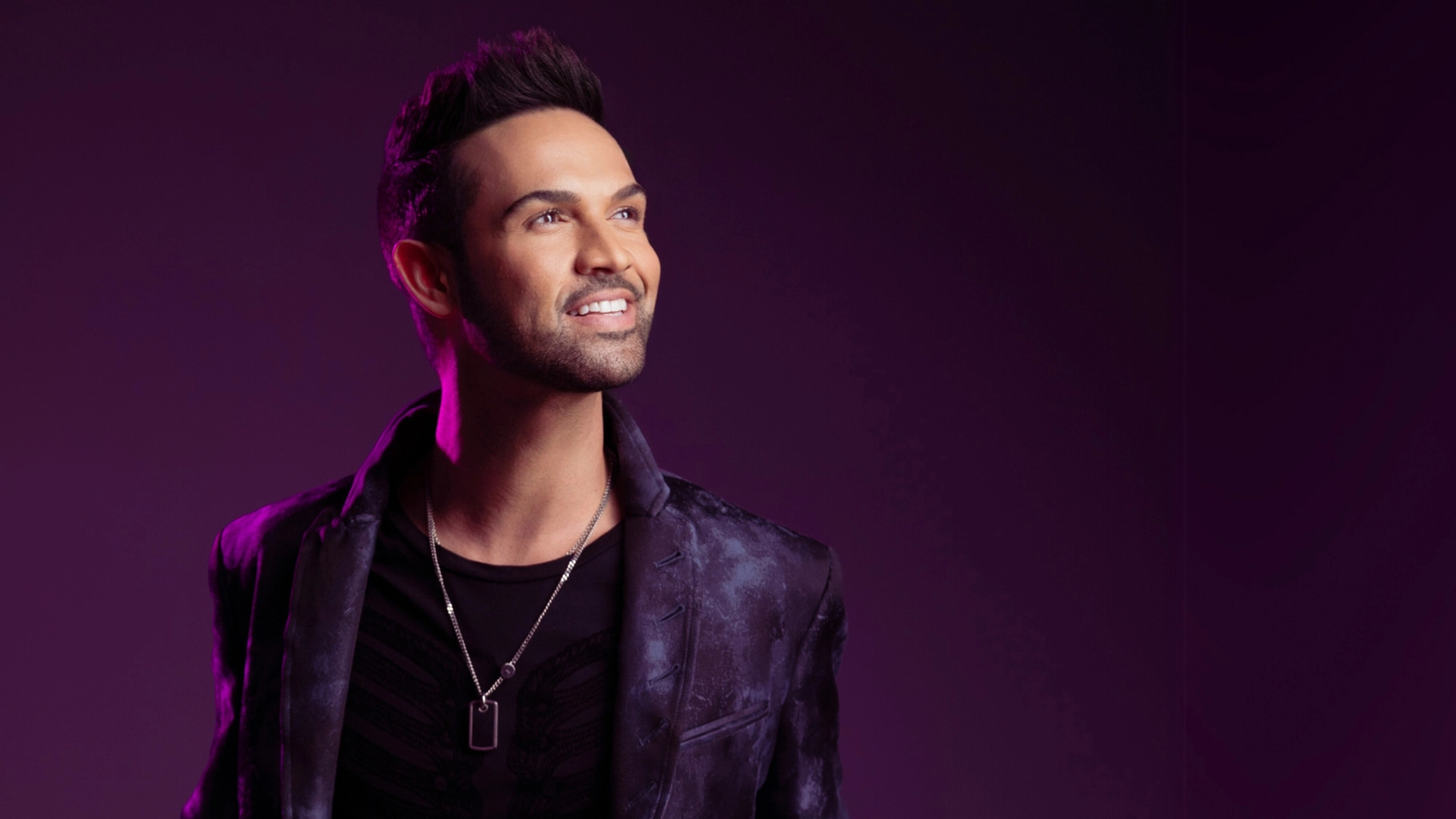 A man with styled dark hair and trimmed beard smiles while looking up, wearing a patterned dark jacket over a black shirt and silver dog tag necklace. The deep purple gradient background hints he’s no stranger to love or music, channeling Chadwick Johnson vibes.