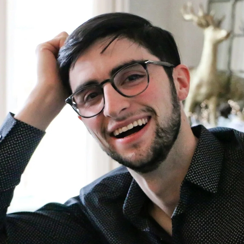 A young man with dark hair, glasses, and a trimmed beard smiles warmly at the camera. He is wearing a dark patterned shirt and rests his head on his hand. In the blurred background, a gold decorative reindeer and an Amanda Quaid poster are visible.