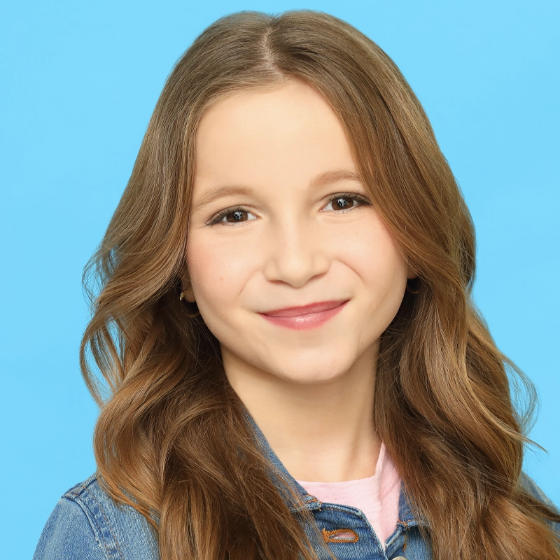 Lia Christina, a young girl with long, wavy light brown hair, smiles gently. She wears a denim jacket over a light pink shirt, set against a solid bright blue background that creates a vibrant and cheerful atmosphere.