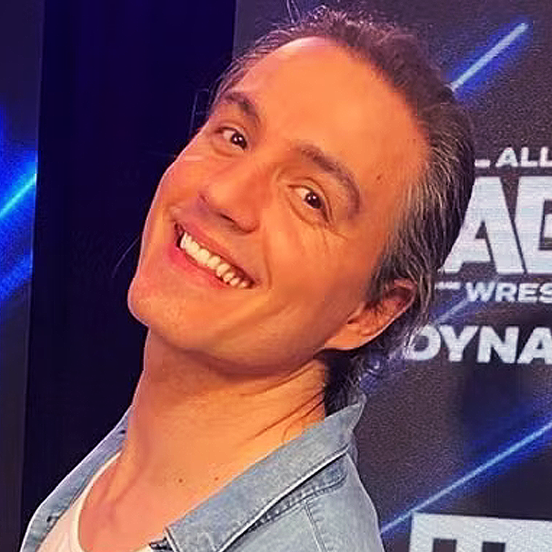 A person with long brown hair pulled back smiles brightly, head tilted to the side. They wear a light blue shirt over a white top. The background is dark with blue lights and partially visible text related to wrestling.