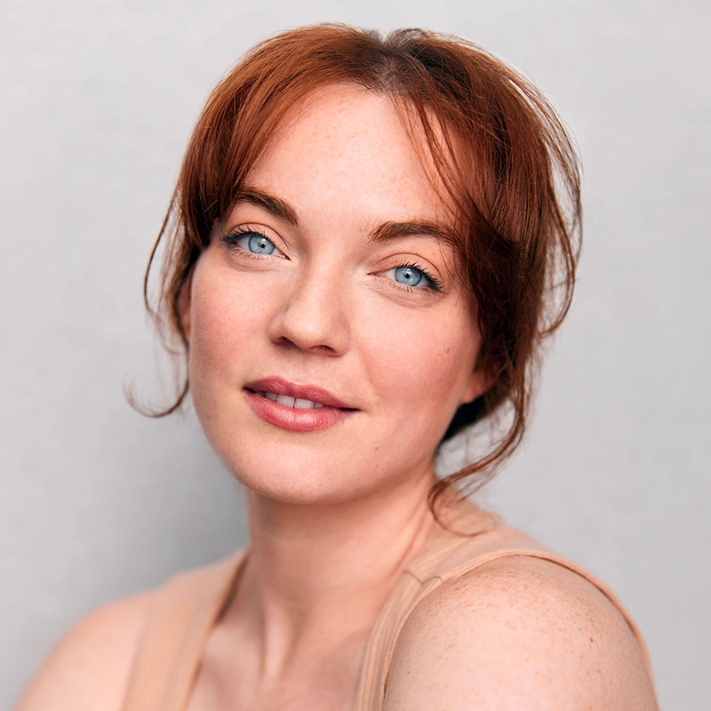 Claire Leyden, a woman with fair skin, blue eyes, and wavy auburn hair, looks at the camera with a slight smile. She wears a sleeveless beige top and minimal makeup against a plain light gray background for a soft, natural look.