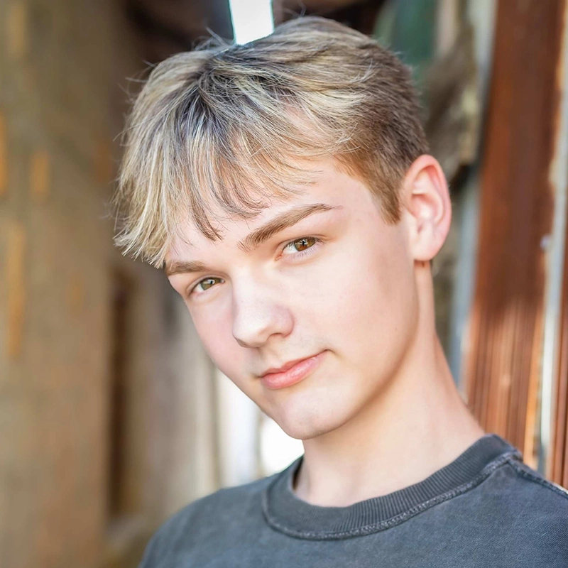 A teenage boy with short, light blonde hair and fair skin looks at the camera with a slight smile. He wears a dark gray t-shirt. The sunlit, outdoor setting hints at Shayla Lopez’s favorite spots—narrow passageways with warm walls.