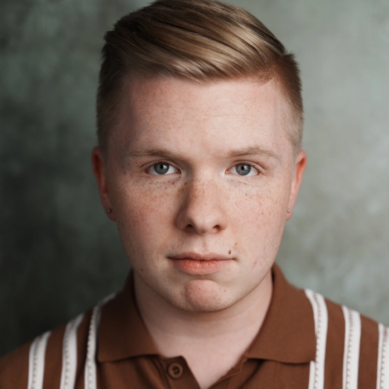Logan Clinger, with short, neatly styled light brown hair and fair skin, stares straight at the camera. They wear a brown collared shirt with white vertical stripes against a softly blurred green and gray background.