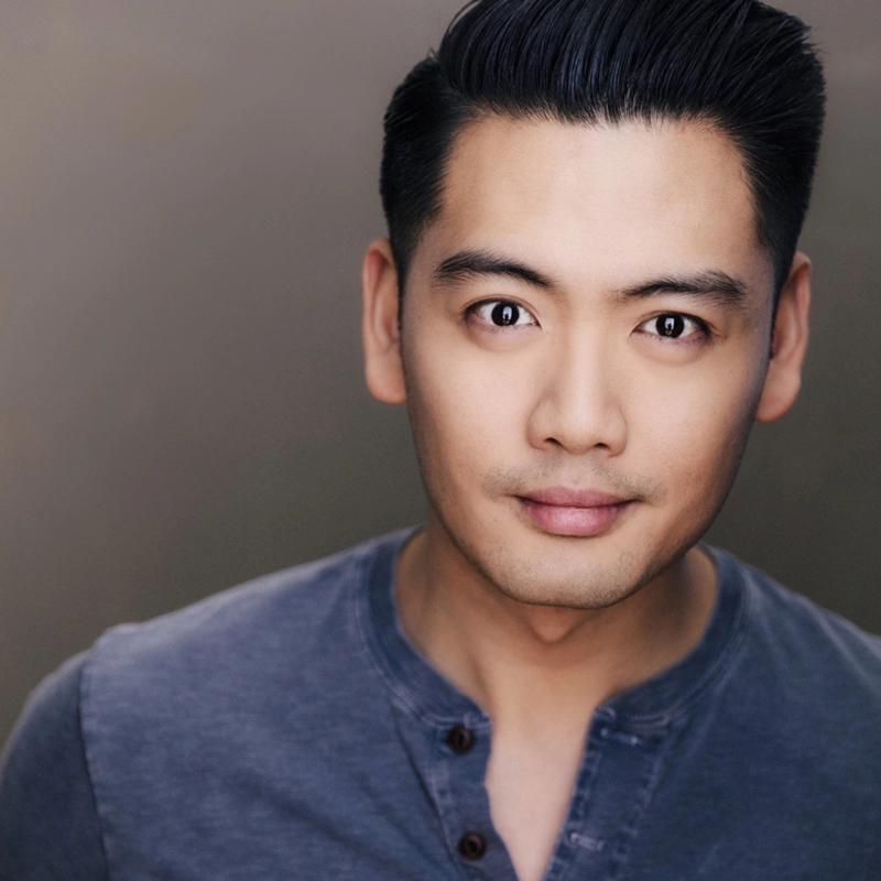 A man with short black hair, styled neatly, looks directly at the camera with a slight smile. Karl Josef Co is wearing a blue, buttonless henley shirt. The softly blurred background in neutral tones highlights his face and expression.