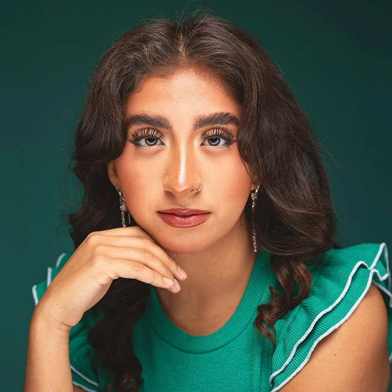 Sophia Leverett, a young woman with long dark hair and bold eyebrows, gazes confidently at the camera. She wears a green top with ruffled sleeves, subtle earrings, and natural makeup. Her hand rests below her chin against a deep green background.