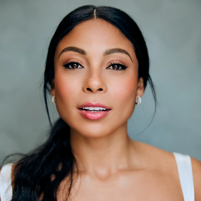Luli Mitchell, a woman with long dark hair pulled back, glowing skin, and natural makeup looks directly at the camera. She wears earrings and a white sleeveless top. The background is softly blurred in shades of gray.