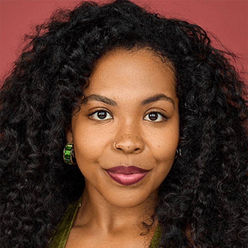 A woman with dark curly hair, medium brown skin, and brown eyes looks directly at the camera. She has a nose ring, wears green hoop earrings, and a dark pink lipstick. The background is a solid mauve color.
