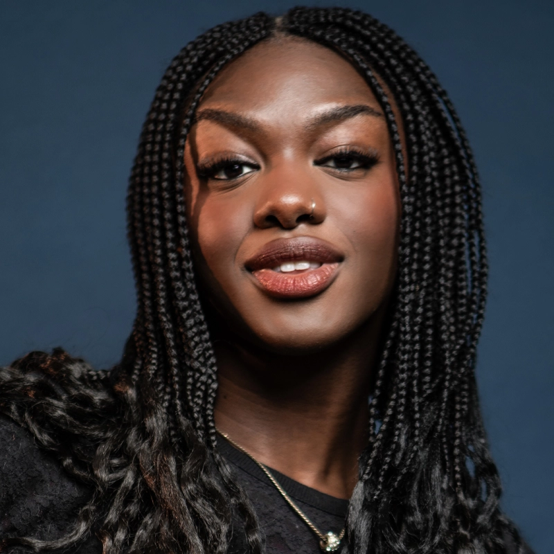 A confident young woman, Destiny David, with long, neat box braids, a nose ring, and natural makeup smiles slightly against a dark blue background. She wears a black top and a delicate necklace. Her expression is friendly and poised.