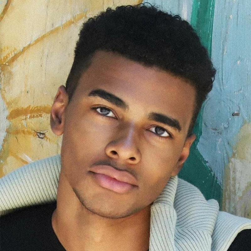 A young man with short curly hair and smooth skin looks into the camera with a neutral expression. He wears a light-colored, textured jacket over a black shirt. The background features distressed wooden panels in beige and teal colors.